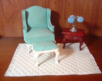 May include: A teal blue armchair with a matching ottoman, a white rug with fringe, and a small table with a lamp with blue glass shades.