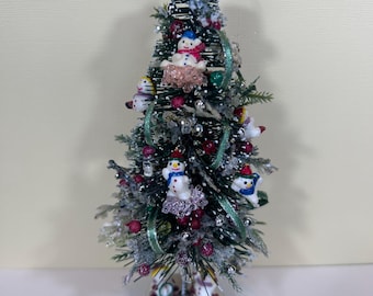 May include: A miniature Christmas tree decorated with snowmen, red berries, and silver accents. The tree is topped with a black hat adorned with a reindeer and red trim. The base is embellished with rhinestones, creating a festive holiday decoration.