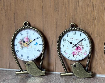 May include: Two bronze-toned metal pendants shaped like clocks with a bird perched on a rope-textured frame. The left clock features Roman numerals and a floral design with a blue butterfly. The right clock displays Arabic numerals and the word Love.