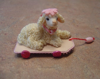 May include: A white lamb with a pink bow and hat is on a pink and white cardboard pull toy. The toy has pink wheels and a red string attached to a pink bead.
