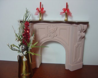 May include: A pink painted miniature fireplace with a decorative carved design and a gold metal candle holder on each side. A vase with a red and white floral arrangement sits in front of the fireplace.