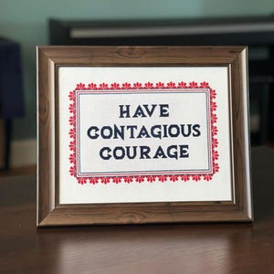 Op de afbeelding: Een ingelijst kruissteekkunstwerk met de woorden "HAVE CONTAGIOUS COURAGE" in marineblauw. De tekst staat gecentreerd op een witte achtergrond, omlijst door een rood-witte bloemenrand, in een houten lijst.