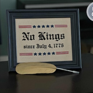 May include: Framed artwork with the text "No Kings since July 4, 1776" in black lettering, with red and blue accents. A gold feather quill rests in front of the frame. The frame is black.