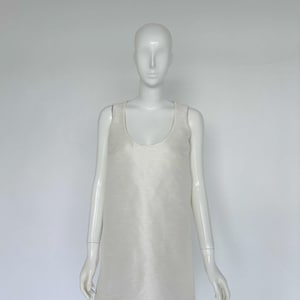 May include: A sleeveless, ivory-colored dress with a scoop neckline. The dress has a simple, A-line silhouette and appears to be made of a smooth, possibly silk-like fabric. The dress is displayed on a mannequin against a plain white background.
