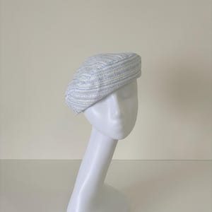 May include: A light blue and white striped beret hat. The hat is displayed on a white mannequin head. The beret has a textured appearance with horizontal stripes. The hat is a stylish accessory.