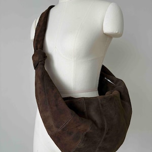 Puede incluir: Un bolso de hombro de ante marrón con forma de media luna. El bolso tiene una correa larga anudada en la parte superior. Se muestra en un maniquí blanco. El bolso tiene un cierre de cremallera.