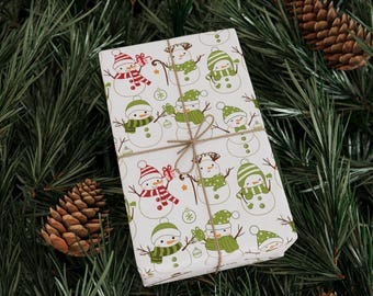 Op de afbeelding: Een ingepakt cadeau met een feestelijk sneeuwpop patroon in rood en groen op een witte achtergrond. Het cadeau is vastgebonden met touw en ligt op een bed van dennennaalden, met dennenappels op de achtergrond.