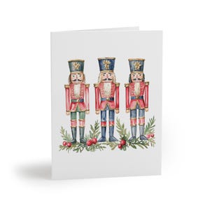 May include: A greeting card featuring three watercolor nutcracker soldiers. Each soldier wears a red coat, blue pants, and a tall hat. They stand on a bed of green leaves and red berries, perfect for the holiday season.