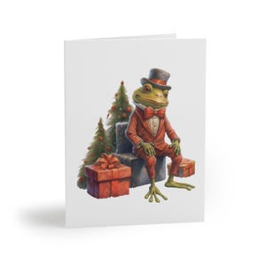 Puede incluir: Una tarjeta de felicitación con una rana elegante con un traje rojo, sombrero de copa y pajarita, sentada entre regalos envueltos y árboles de Navidad. La rana tiene piel verde y está sentada en un bloque de piedra. La tarjeta es blanca.