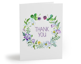 Könnte beinhalten: Eine weiße, gefaltete Grußkarte mit einem Aquarell-Blumenkranz. Der Kranz zeigt lila, gelbe und weiße Blumen, grüne Blätter und die Worte "THANK YOU" in Lila. Die Karte ist innen leer.