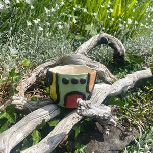 May include: A small, hand-painted ceramic planter with a geometric design in green, yellow, orange, and red. The planter sits on a weathered, gray branch, surrounded by greenery and small white flowers. The planter is approximately 10 cm wide.