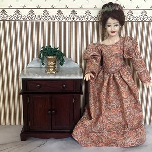 May include: A miniature doll in a patterned dress with a floral crown stands next to a small wooden cabinet. The cabinet has a marble top and a gold-colored vase with greenery. The doll's dress features a repeating floral pattern in shades of peach and brown.