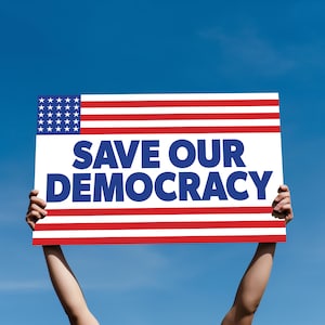 May include: A person holds a sign with the American flag in the top left corner and the words "SAVE OUR DEMOCRACY" in blue letters on a white background.