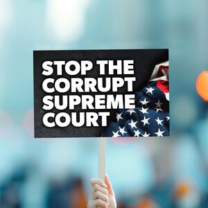 May include: A sign held up with the words "STOP THE CORRUPT SUPREME COURT" in bold white letters. The sign is held up by a wooden stick. The American flag is visible in the background.