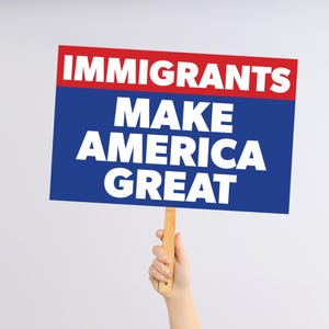 以下が含まれることがあります： 「IMMIGRANTS MAKE AMERICA GREAT」の文字が白の太字で青い背景に書かれた看板を人が持っています。上部には赤いバナーがあります。看板は木の棒で支えられています。