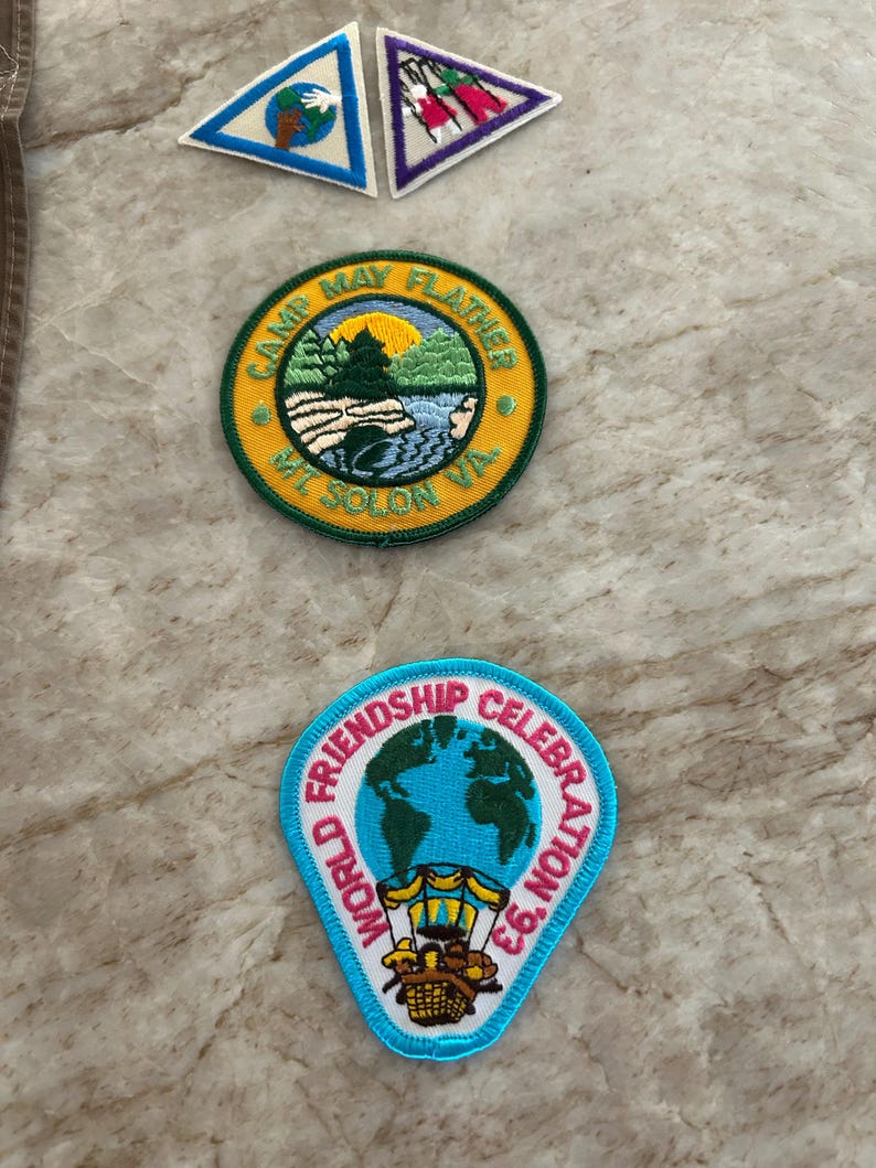 May include: A collection of embroidered patches. Includes two triangular patches with nature scenes and a round patch with "Camp May Flather Mt. Solon VA" text and a landscape. A blue and white patch reads "World Friendship Celebration '93".