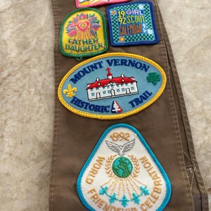 May include: Brown fabric with several embroidered patches. Patches include text: "BRIDGING", "FATHER DAUGHTER", "19 GIRL 92 SCOUT CALENDAR", "MOUNT VERNON HISTORIC TRAIL", and "WORLD FRIENDSHIP CELEBRATION 1992".