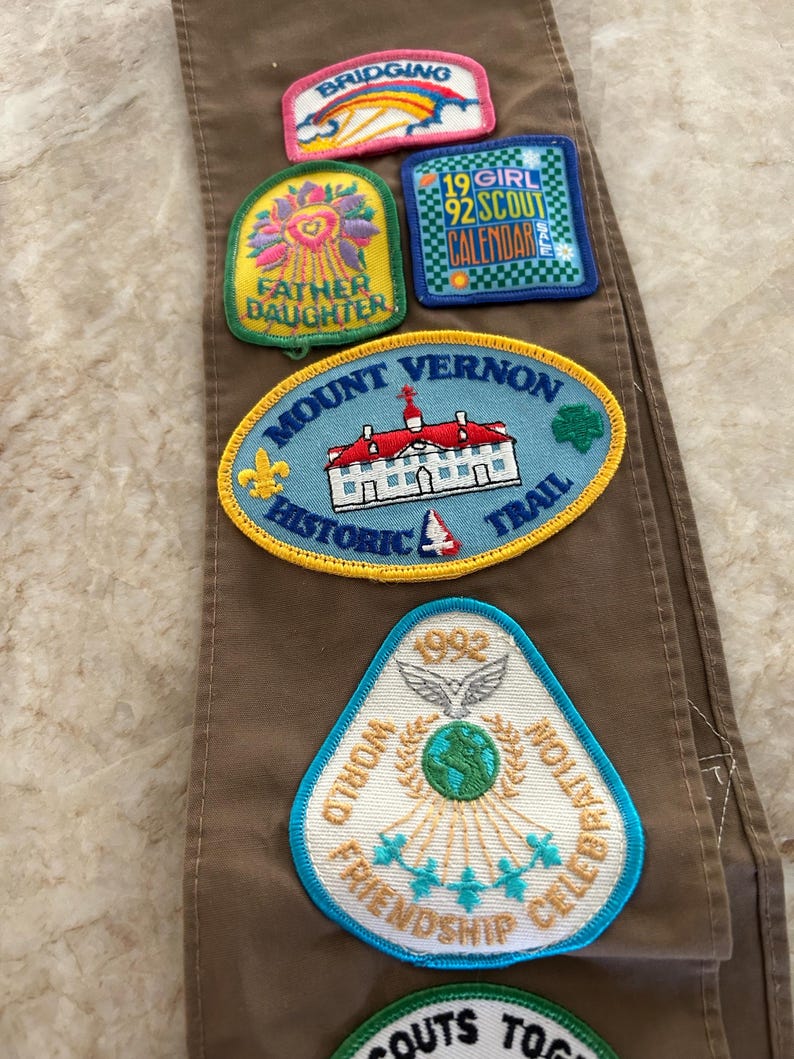 May include: Brown fabric with several embroidered patches. Patches include text: "BRIDGING", "FATHER DAUGHTER", "19 GIRL 92 SCOUT CALENDAR", "MOUNT VERNON HISTORIC TRAIL", and "WORLD FRIENDSHIP CELEBRATION 1992".