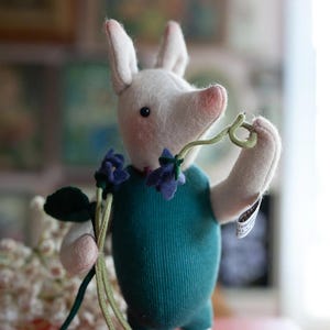 May include: A handmade plush pig wearing a teal outfit and holding a sprig of flowers. The pig has a white body, pink snout and ears, and is adorned with a blue flower necklace. The toy is standing on a red surface.