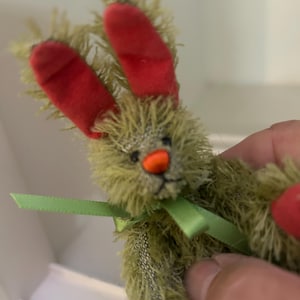 May include: A small, handmade plush bunny with olive green fur and red velvet ears. It has a small orange nose and a green ribbon tied around its neck. The bunny is a whimsical, handcrafted item.