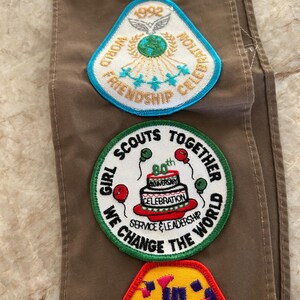 May include: A collection of colorful embroidered patches, including a blue and yellow "Historic Trail" patch, a white and blue "World Friendship Celebration" patch, and a green "Girl Scouts Together" patch celebrating an anniversary.