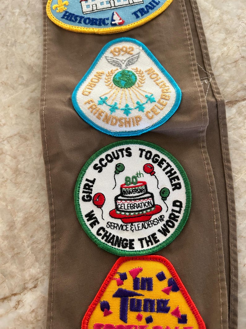 May include: A collection of colorful embroidered patches, including a blue and yellow "Historic Trail" patch, a white and blue "World Friendship Celebration" patch, and a green "Girl Scouts Together" patch celebrating an anniversary.