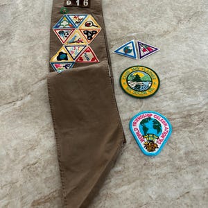 May include: A brown Girl Scout sash with various patches, including a "Friendship Celebration '93" patch and a "Camp May Flather" patch. The sash also features a "916" number and several triangular patches.