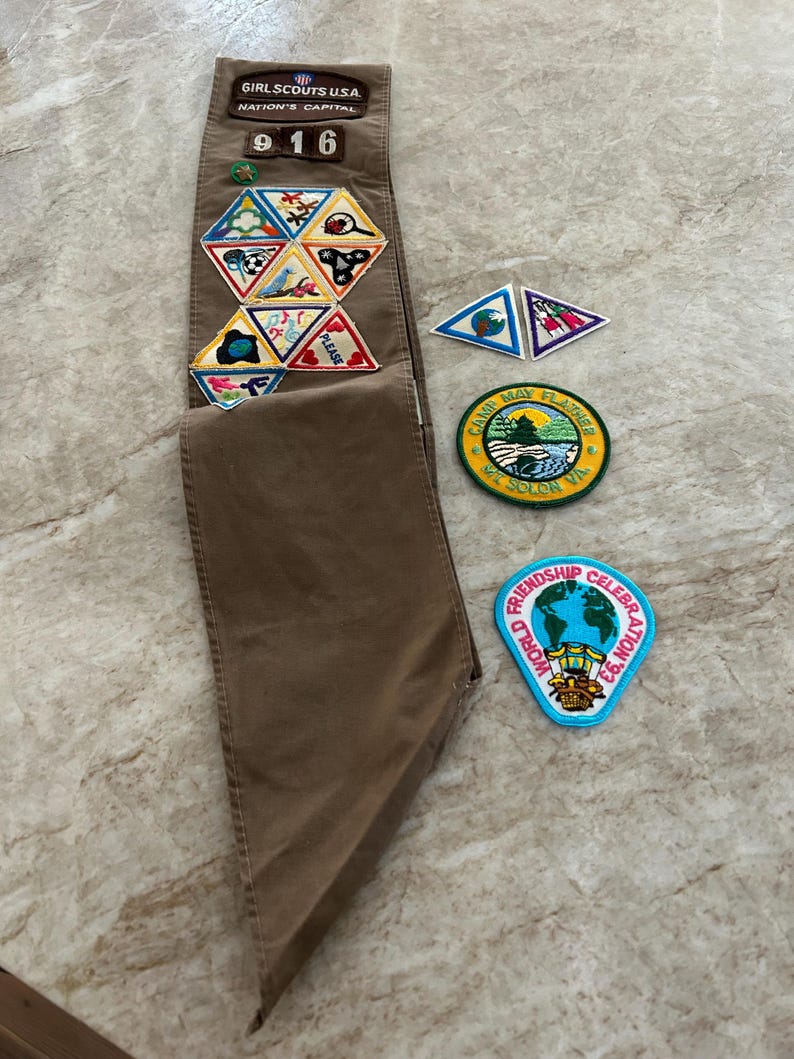 May include: A brown Girl Scout sash with various patches, including a "Friendship Celebration '93" patch and a "Camp May Flather" patch. The sash also features a "916" number and several triangular patches.