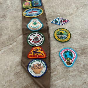 May include: A brown Girl Scout sash with various colorful patches. Patches include text like "Mount Vernon Historic Trail", "Friendship Celebration", and "Cookie Sale". Other patches feature images of a bear, a cake, and a globe.