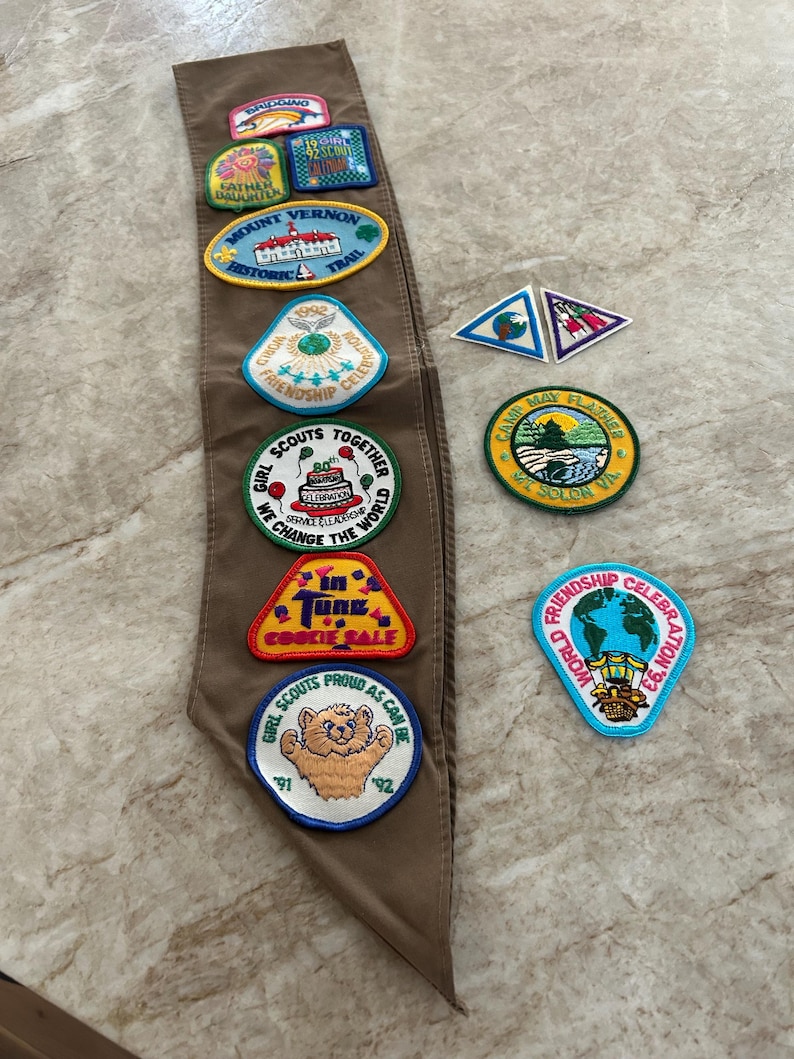 May include: A brown Girl Scout sash with various colorful patches. Patches include text like "Mount Vernon Historic Trail", "Friendship Celebration", and "Cookie Sale". Other patches feature images of a bear, a cake, and a globe.