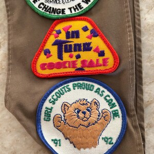 May include: Three embroidered Girl Scout patches on a tan fabric. The patches include text and images: "We Change the World," "In Tune Cookie Sale," and a bear with "Girl Scouts Proud as Can Be" and the years '91 and '92.