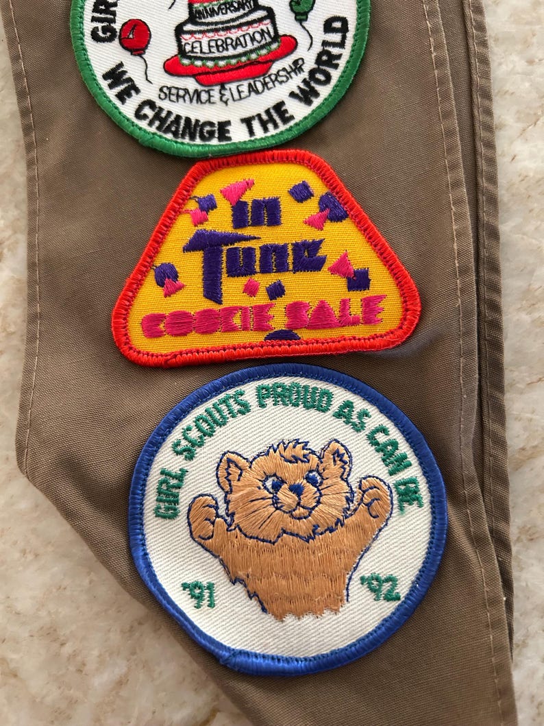 May include: Three embroidered Girl Scout patches on a tan fabric. The patches include text and images: "We Change the World," "In Tune Cookie Sale," and a bear with "Girl Scouts Proud as Can Be" and the years '91 and '92.