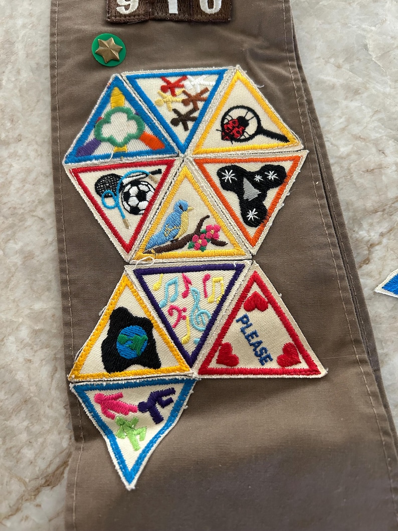 May include: Brown sash with embroidered patches. Patches include a soccer ball, a blue bird, musical notes, and the word "PLEASE". A green and gold star-shaped pin is also visible.