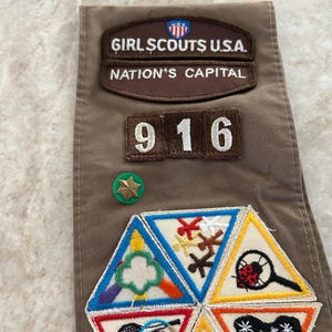 May include: A brown Girl Scouts sash with patches and pins. Patches include "Girl Scouts U.S.A. Nation's Capital" and the number "916". Additional patches feature various activity symbols. A green and gold star pin is also visible.