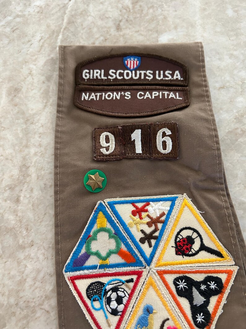 May include: A brown Girl Scouts sash with patches and pins. Patches include "Girl Scouts U.S.A. Nation's Capital" and the number "916". Additional patches feature various activity symbols. A green and gold star pin is also visible.