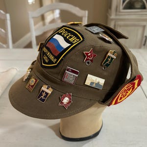 May include: A brown military style cap with a red star and the word "Россия" (Russia) on the front. The cap is decorated with various pins and patches, including a red star, a hammer and sickle, and a running figure.