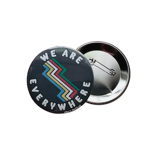 May include: A round button with a black background and white text that reads "We are everywhere". The text is in a circle around a colorful, abstract design. The button has a silver back with a safety pin.