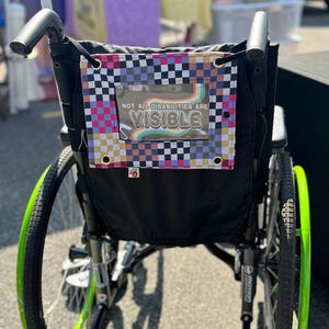 May include: A black wheelchair with a green rimmed wheel. The back of the wheelchair has a black fabric pouch with a checkered pattern in pink, blue, yellow, and white. The pouch has a clear plastic window with the text "NOT ALL DISABILITIES ARE VISIBLE" in rainbow letters.