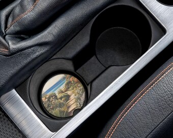 May include: A car cup holder with a round coaster featuring a coastal landscape scene. The coaster has a light blue sky, ocean, and a path with plants. The cup holder is black and sits in a silver and black car console.