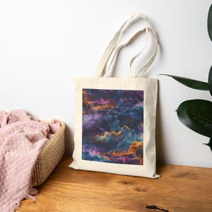 May include: A cream-coloured tote bag displaying a nebula design. The image shows swirling clouds in shades of purple, blue, and orange, dotted with small white stars. The bag is placed on a wooden surface.