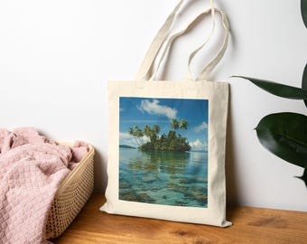May include: A natural-coloured tote bag displaying a tropical island scene. The image depicts a small island with palm trees, surrounded by clear turquoise water under a blue sky with clouds. The bag has long handles.