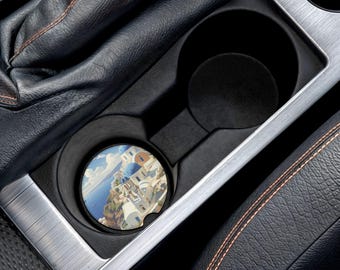 May include: A car cup holder coaster featuring a scenic view of a coastal town with white buildings and blue domes. The coaster is round and fits inside a car's cup holder. The image shows a car's interior with black leather and silver trim.