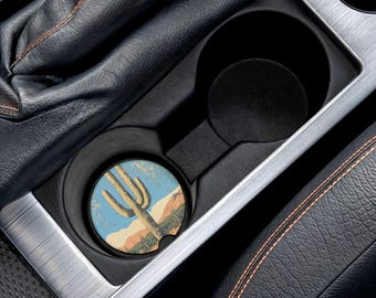 Puede incluir: Un posavasos redondo para coche con un paisaje desértico con un cactus saguaro, montañas y cielo azul. El posavasos está colocado en el portavasos de un coche, rodeado de cuero negro y molduras plateadas. El posavasos tiene un diámetro de aproximadamente 6,35 cm.