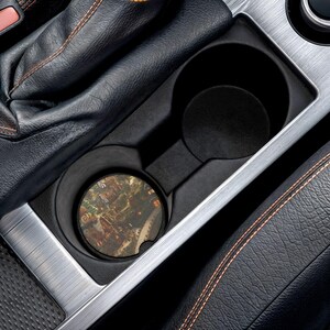 Könnte beinhalten: Ein Getränkehalter im Auto mit einem Untersetzer mit einem detaillierten Stadtbilddesign. Der runde Untersetzer passt in den Getränkehalter, der Teil der Innenkonsole des Autos ist. Der Innenraum ist schwarz mit silbernen Zierleisten und Lederakzenten.