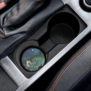 May include: A car cup holder coaster with a whimsical illustration of a small house in a starry night scene. The coaster is round and fits inside a car's cup holder. The image is set against a black interior.