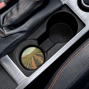 Puede incluir: Un portavasos de coche con un posavasos redondo con una pintura de paisaje de un campo con hileras de cultivos. El posavasos está colocado dentro de un portavasos negro, rodeado de un borde plateado y un interior de cuero negro.