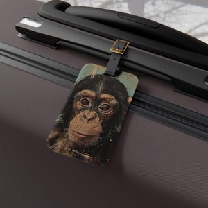 May include: A luggage tag featuring a chimpanzee's face. The tag has a black leather strap and a gold-colored buckle. The background of the tag is a distressed teal color with gold flecks. The tag is attached to a dark gray suitcase.