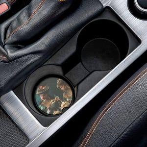 May include: A car cup holder featuring a round coaster with a vintage painting of two women. The coaster has a black border and is set inside a black cup holder. The car's interior is black with silver and orange accents.