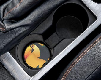 May include: A round car coaster featuring a toucan design in yellow, black, and orange. The coaster is placed in a black car cup holder, surrounded by black leather and silver trim.