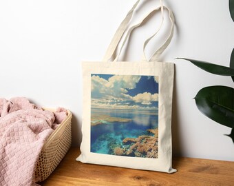 May include: A natural-coloured tote bag displaying a scenic ocean view. The image shows a blue sky with white clouds, clear turquoise water, coral reefs, and a distant shoreline. The bag has long handles and is made of a natural fabric.
